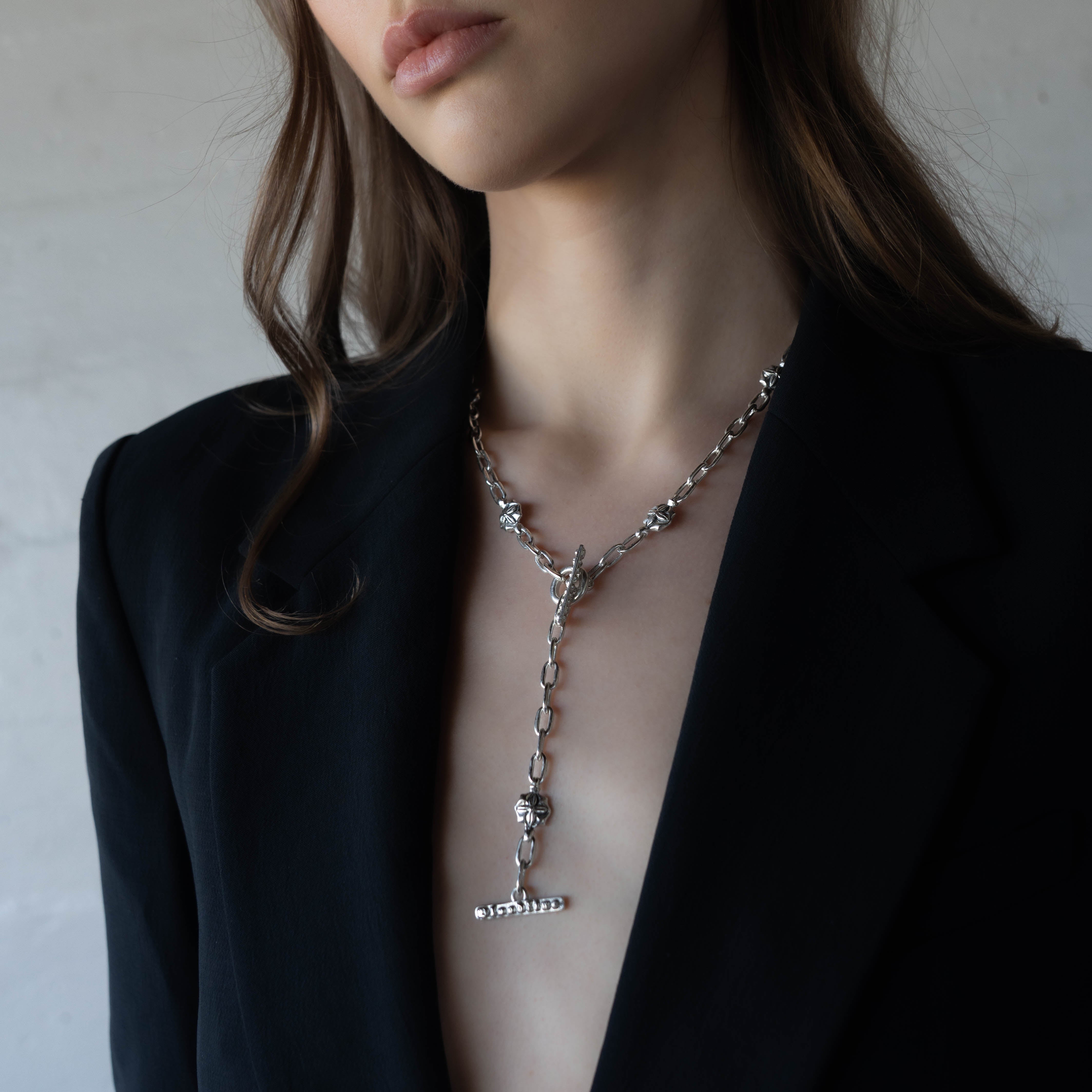 A close-up of a model wearing a silver lariat necklace with a floral links and a T-bar dangle, styled with a black blazer.