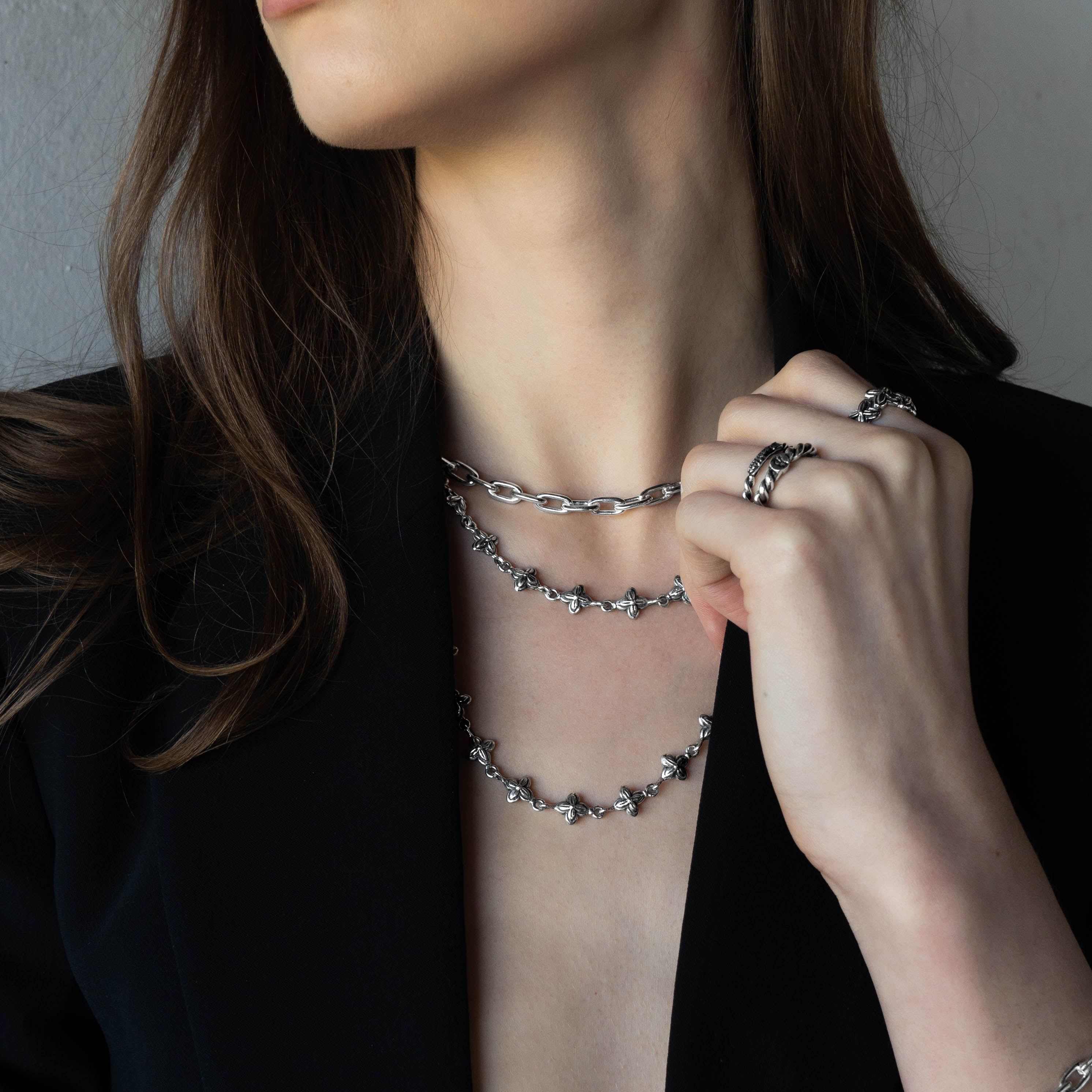 A close-up of a woman wearing layered silver chain necklaces and rings, with her hand touching the jewelry. She is wearing a black blazer against a neutral gray background.