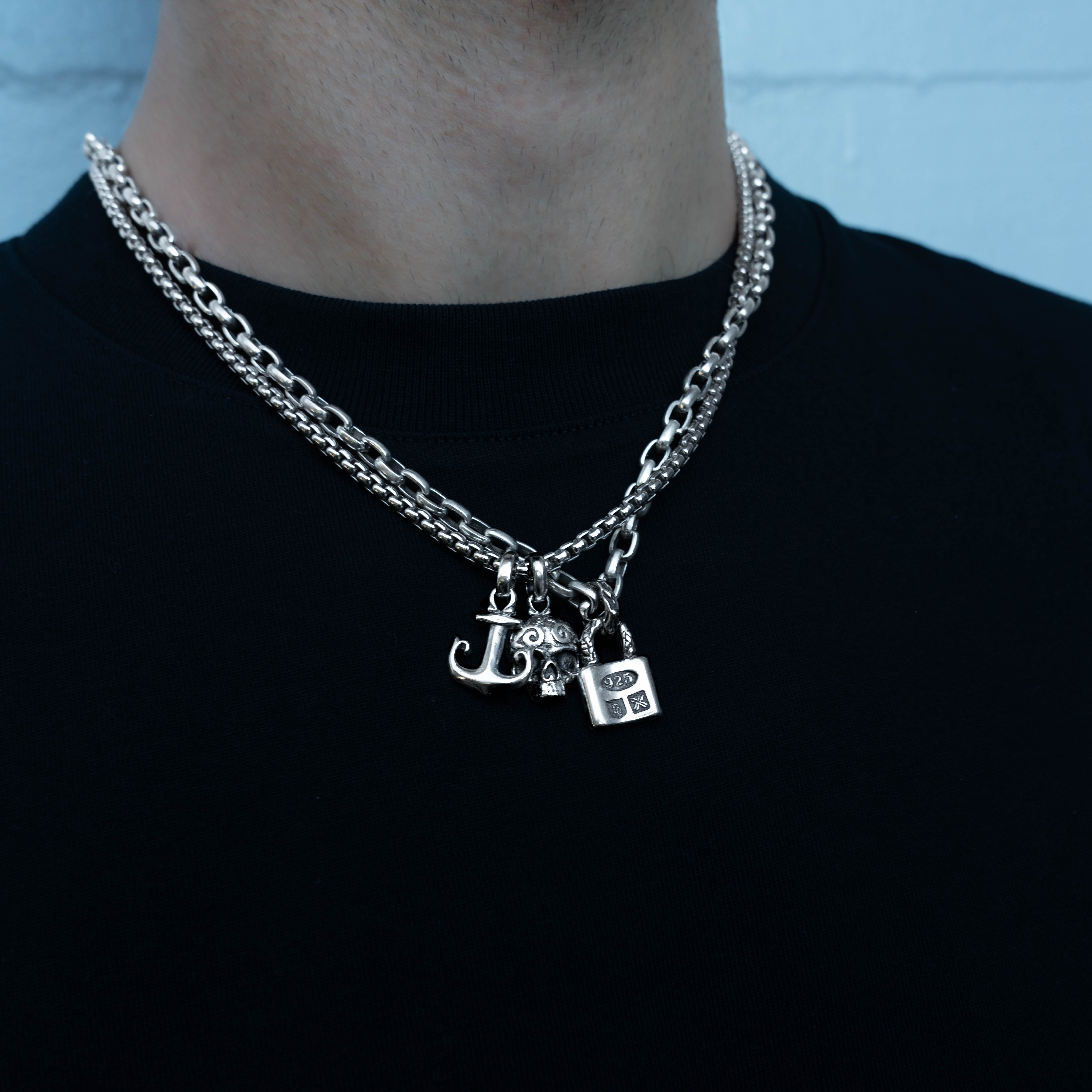 A close-up of a person wearing a layered silver chain necklace with three small charms: an anchor pendant, a sugar skull pendant and a locket with detailed design.