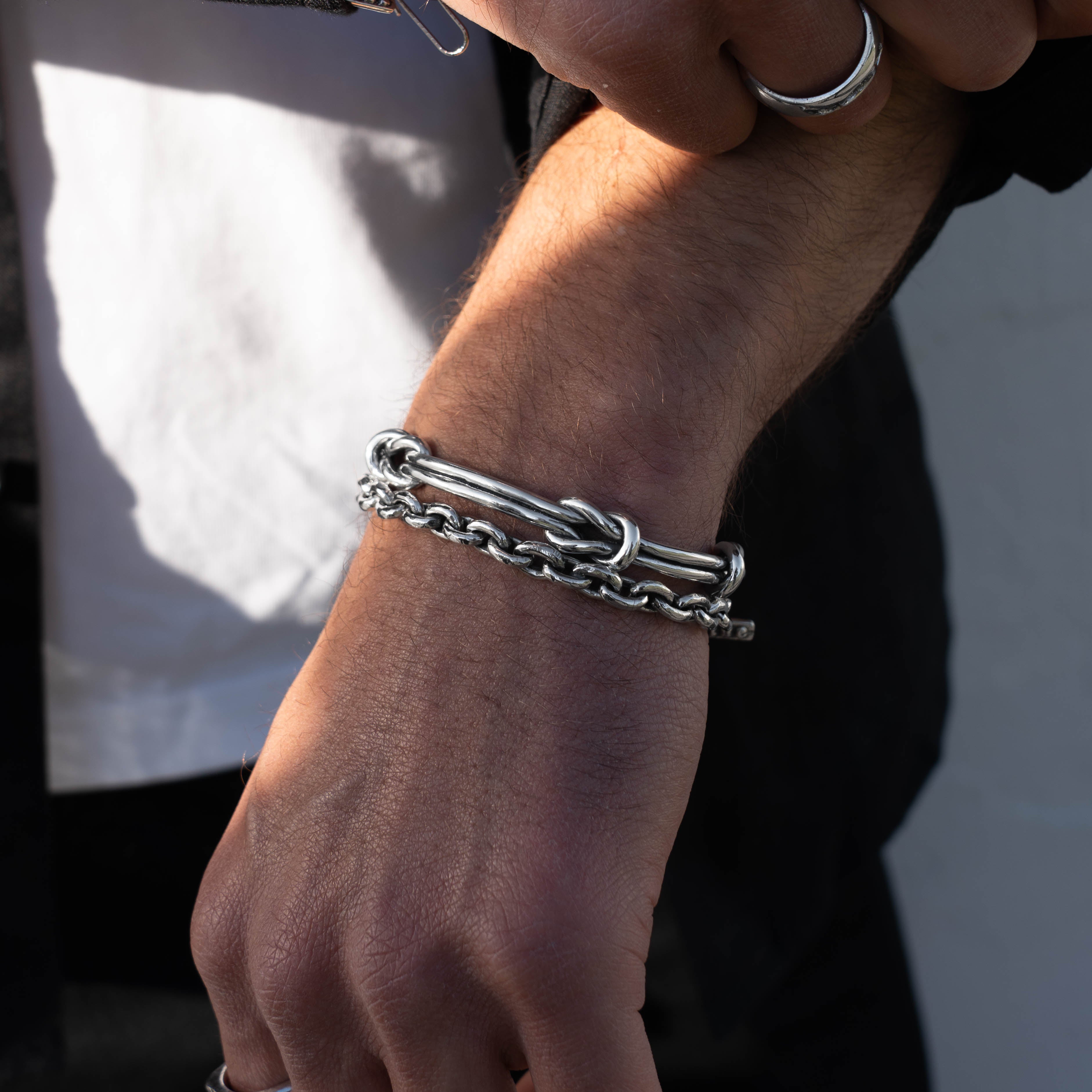 A close-up of a person's wrist wearing a silver chain bracelet with a knotted bar detail, paired with a silver ring on another finger.