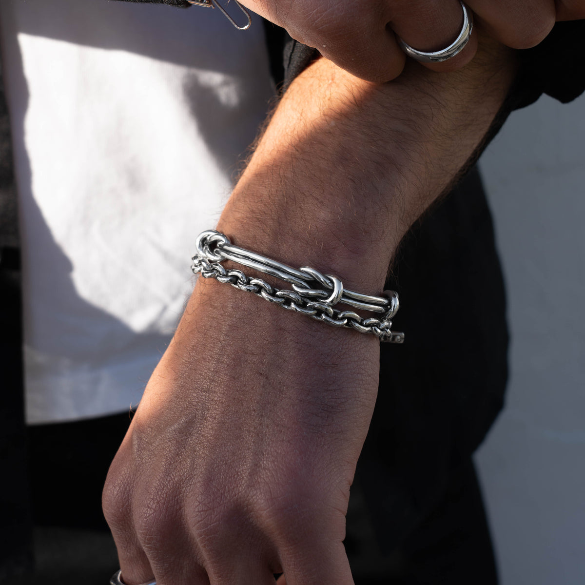 A close-up of a person's wrist wearing a silver chain bracelet with a knotted bar detail, paired with a silver ring on another finger.