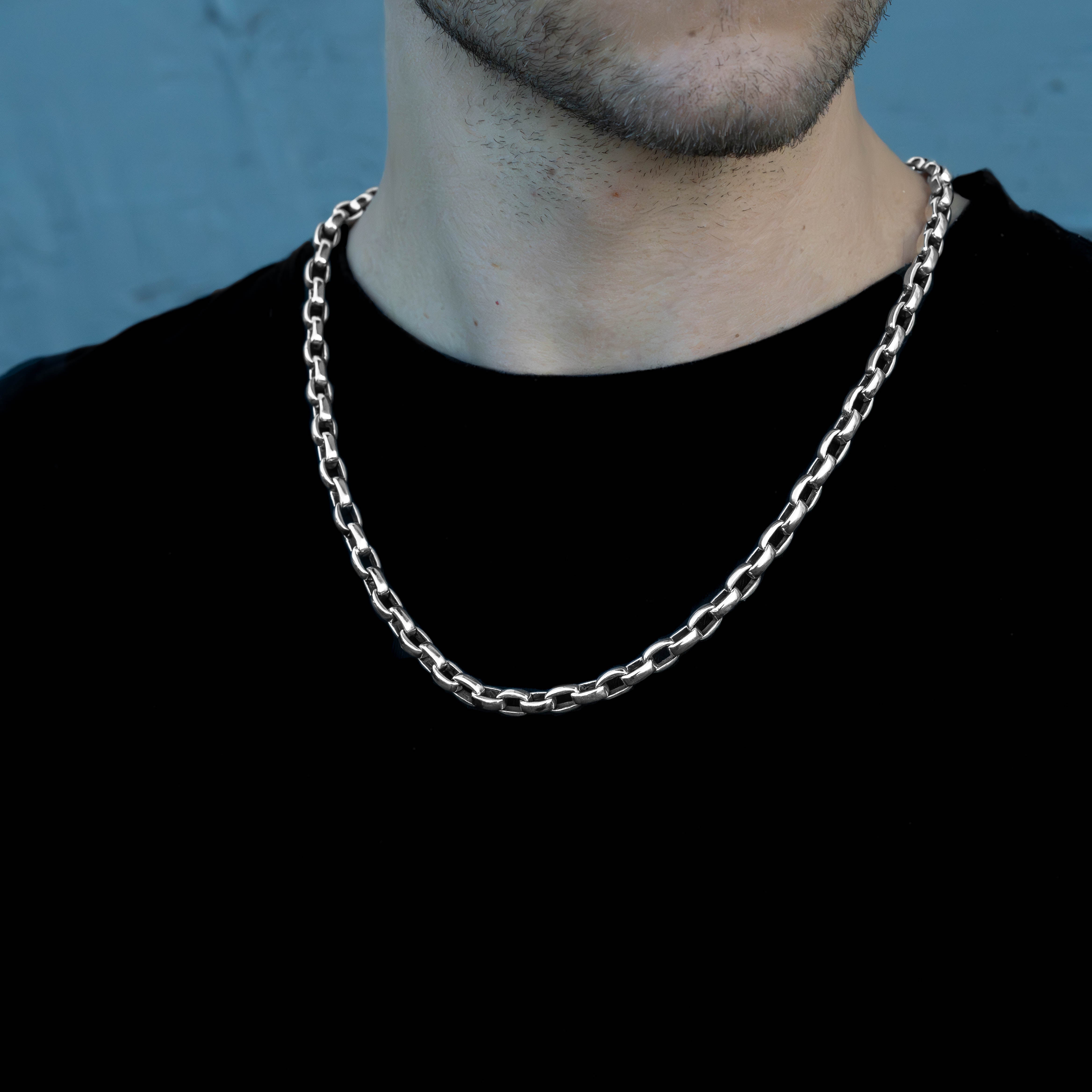 A close-up of a man wearing a thick, polished silver chain necklace over a black t-shirt, set against a textured blue-gray background.
