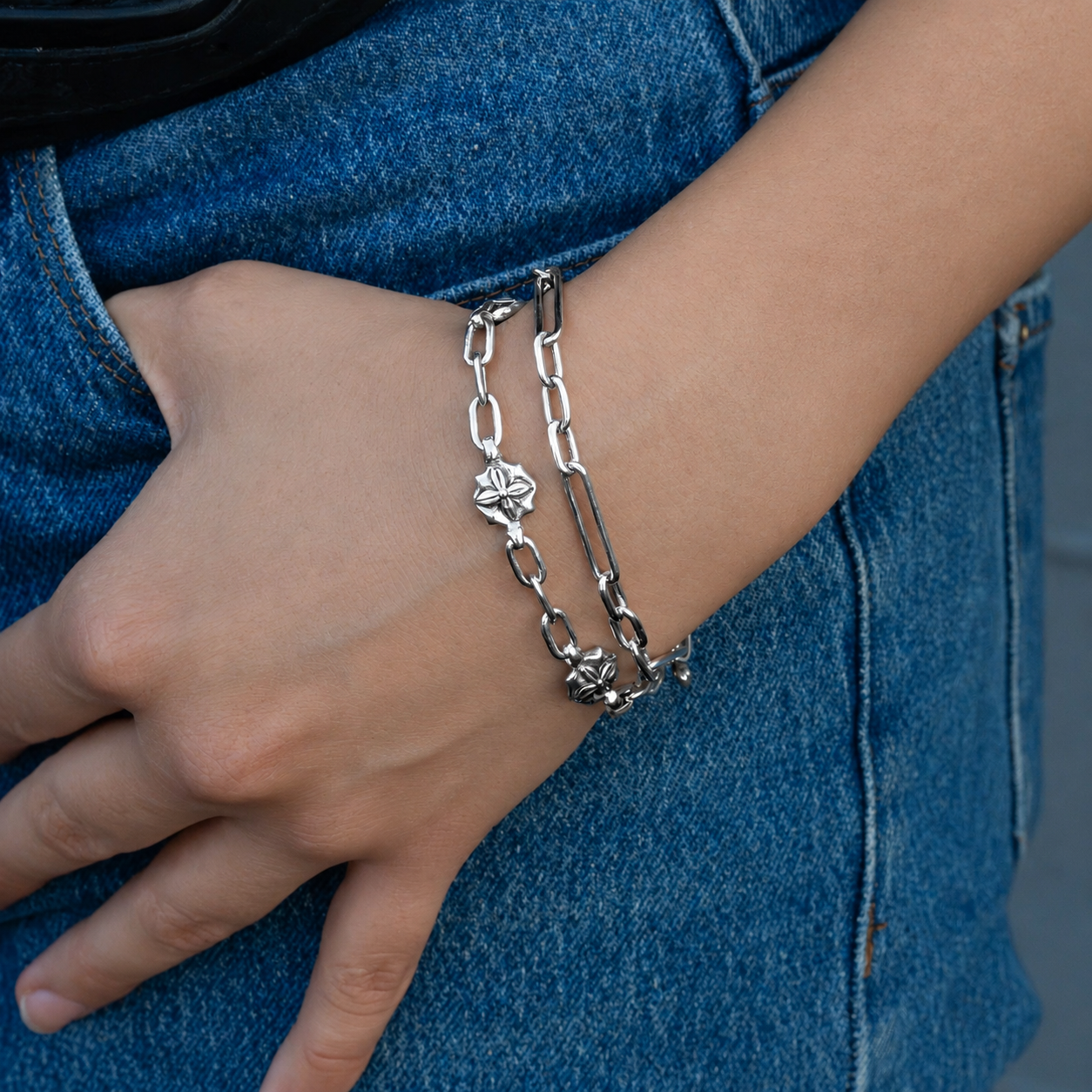 A close-up of a person's wrist wearing two silver chain bracelets, one with a floral charm and one with a flower charm, paired with blue denim jeans.