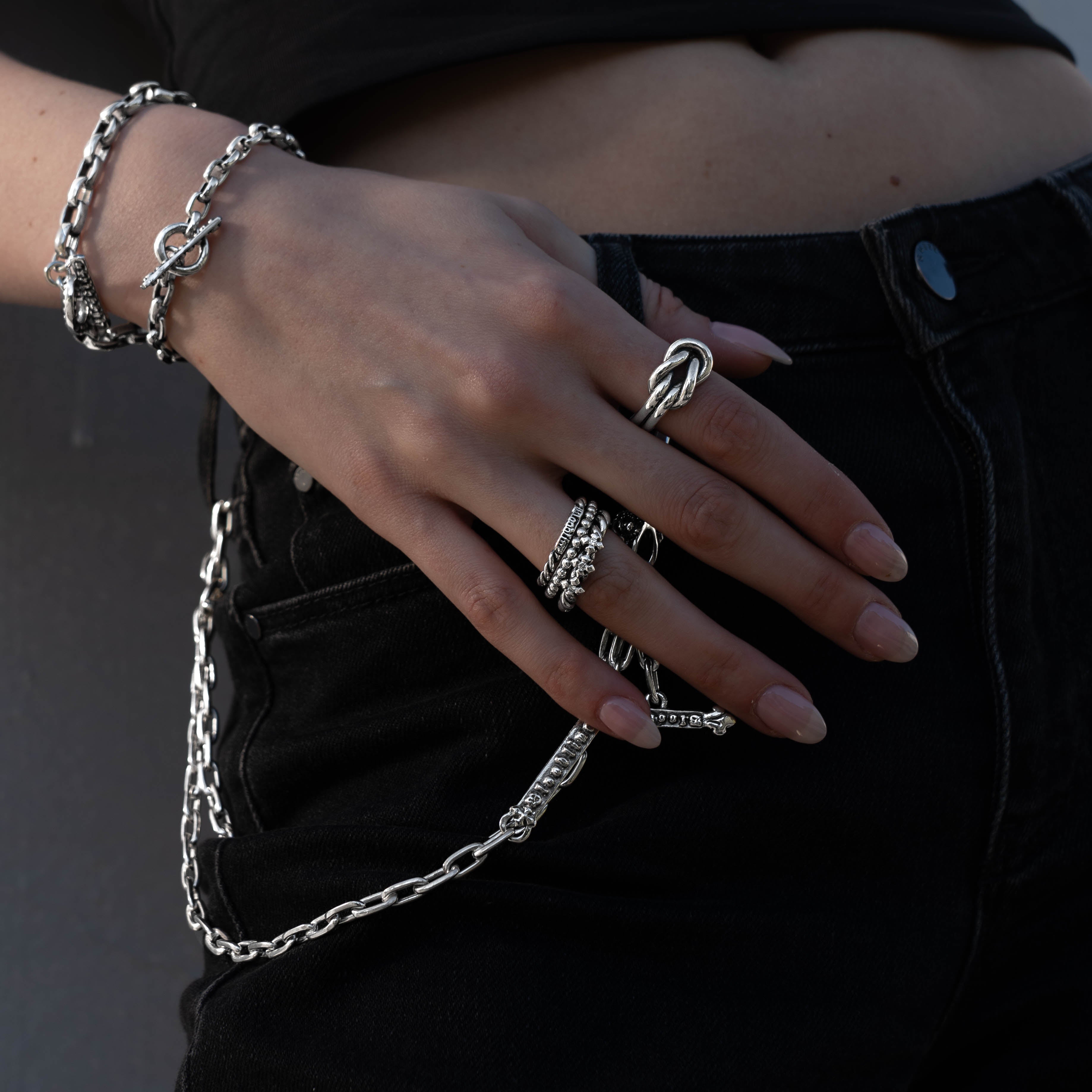 A close-up of a person's hand wearing multiple silver chain-link rings and bracelets, styled with black denim jeans.