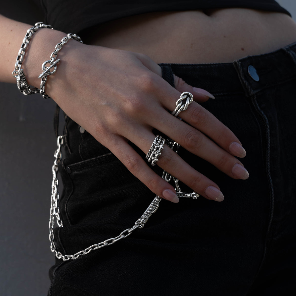 A close-up of a person's hand wearing multiple silver chain-link rings and bracelets, styled with black denim jeans.