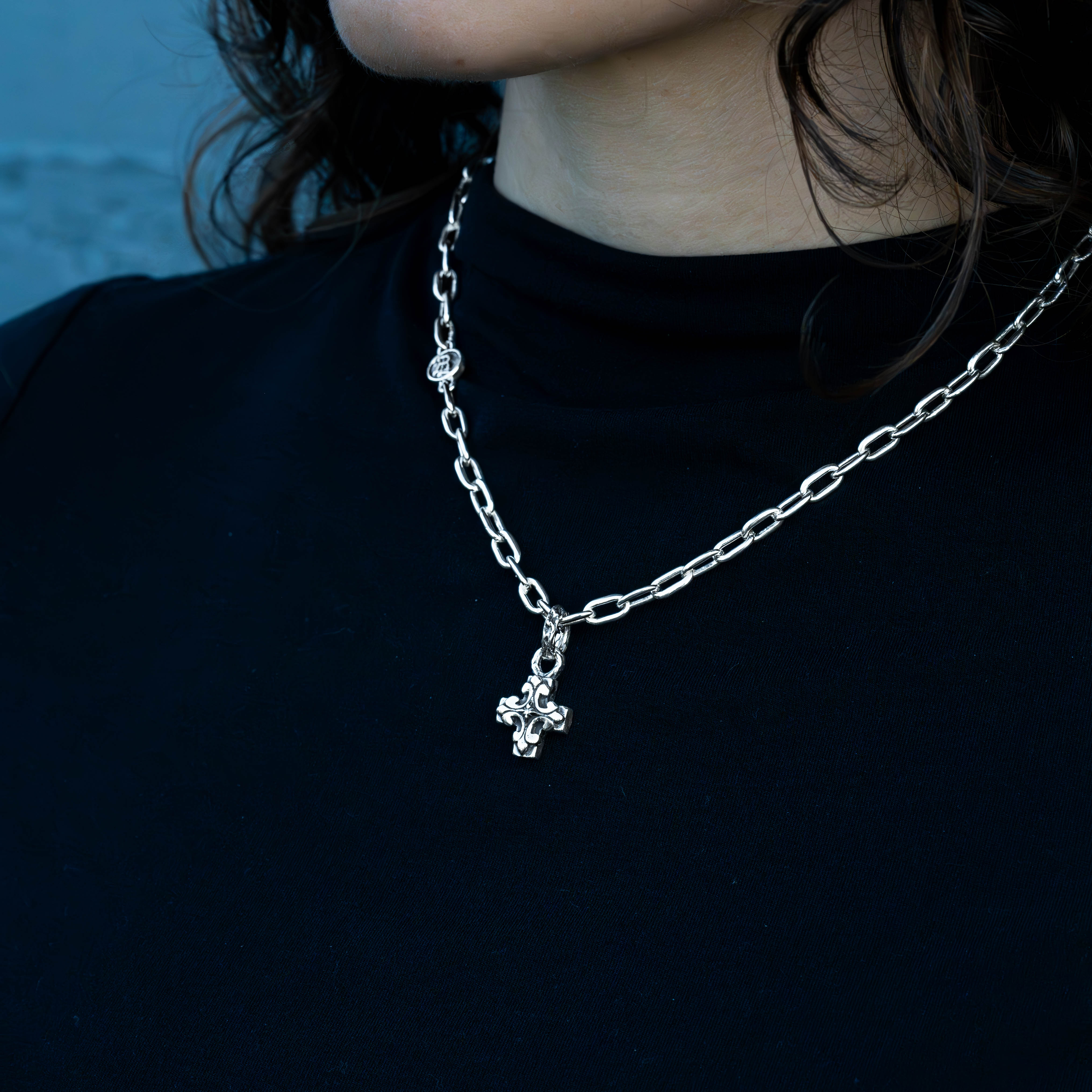 A close-up of a person wearing a silver chain necklace with a small cross pendant, styled with a black t-shirt against a dark blue background.
