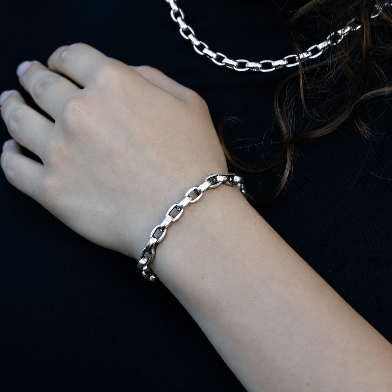 A close-up of a person's wrist wearing a polished silver chain link bracelet, with a matching chain necklace draped near the shoulder against a dark background.