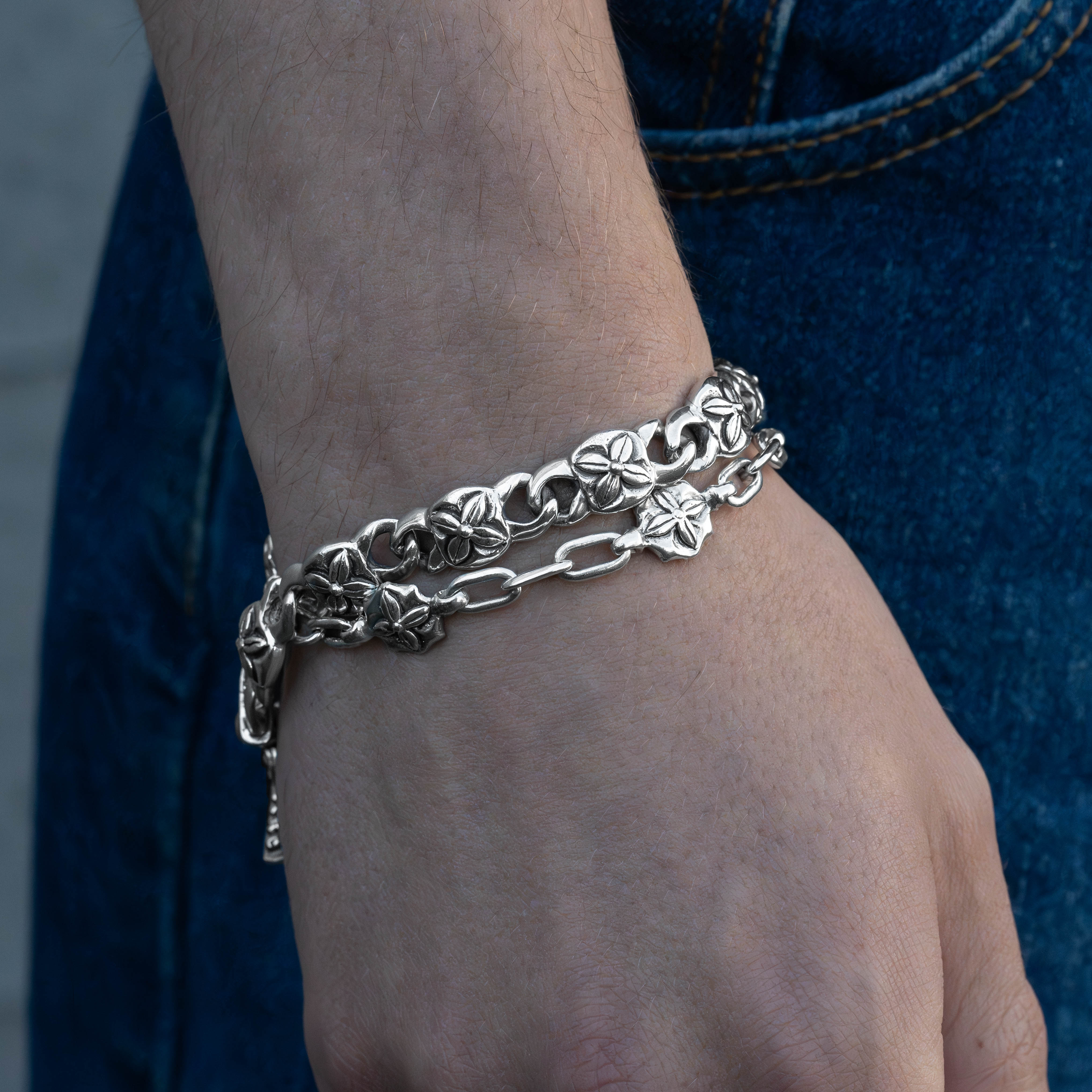 A close-up of a person's wrist wearing a double-layered silver chain bracelet, featuring one layer with textured floral links and the other with smooth oval links, paired with blue denim jeans.
