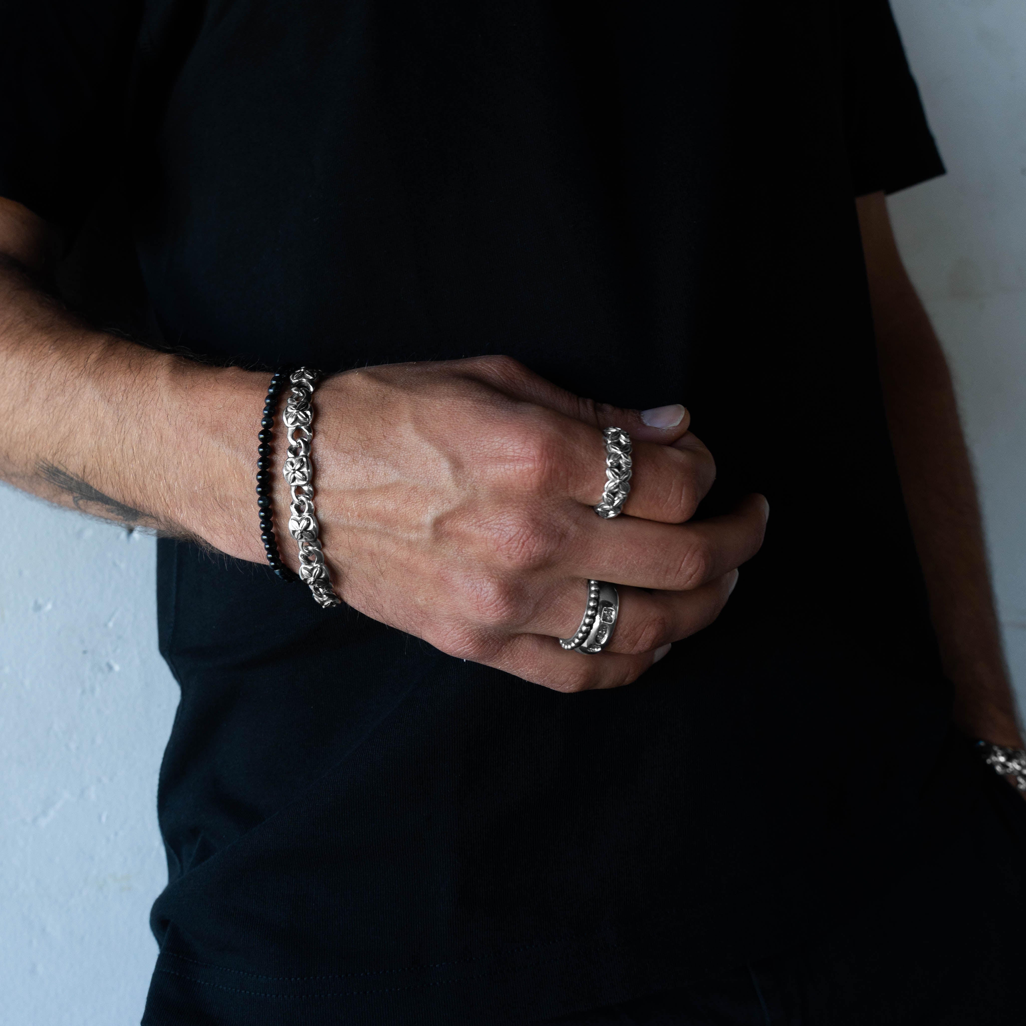 A close-up of a person's hand wearing multiple silver-toned rings and a silver-toned beaded bracelet, dressed in a black t-shirt against a light gray wall.