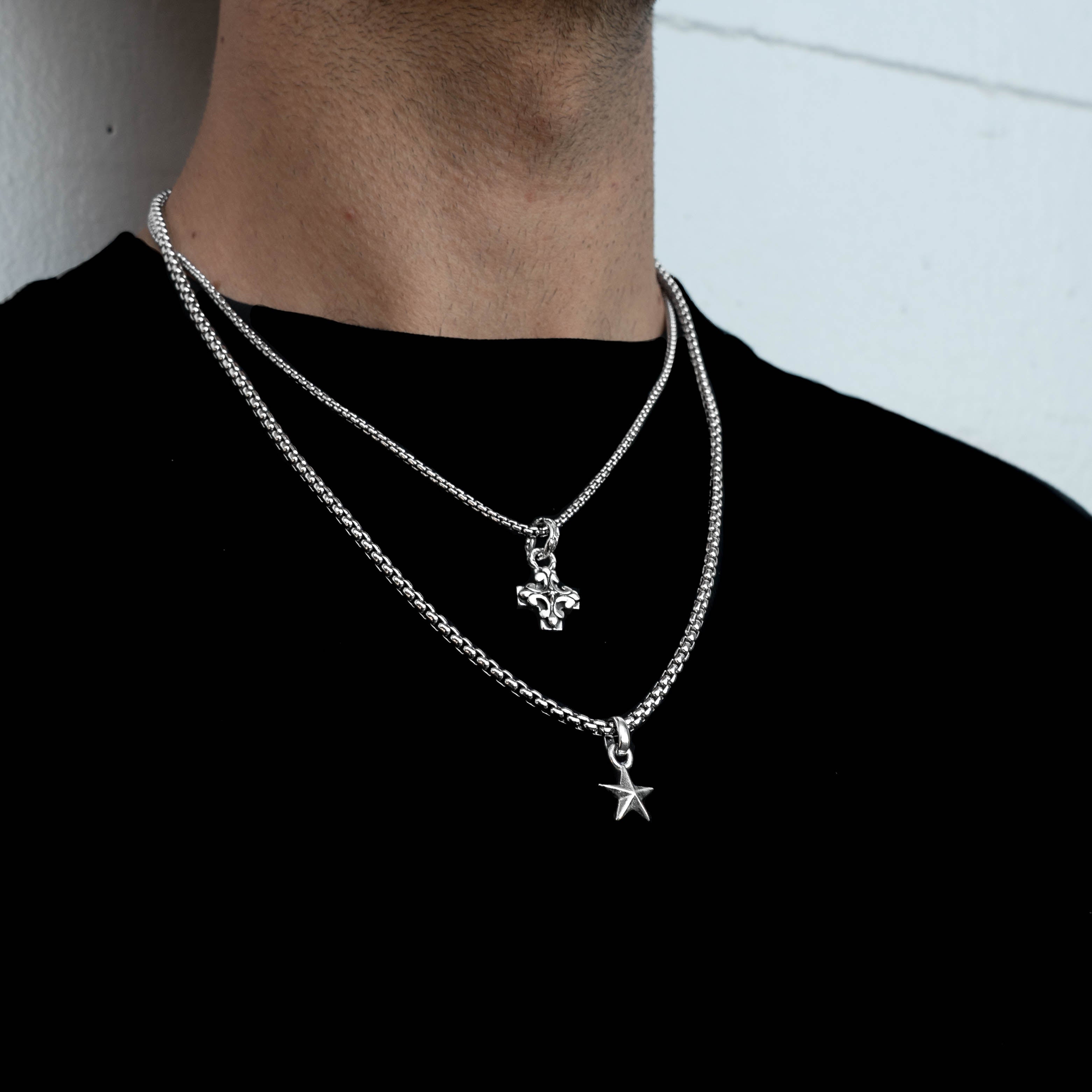 A close-up of a person's neck and upper chest wearing two layered silver chain necklaces. The top chain has a small cross pendant, and the bottom chain has a star-shaped pendant.