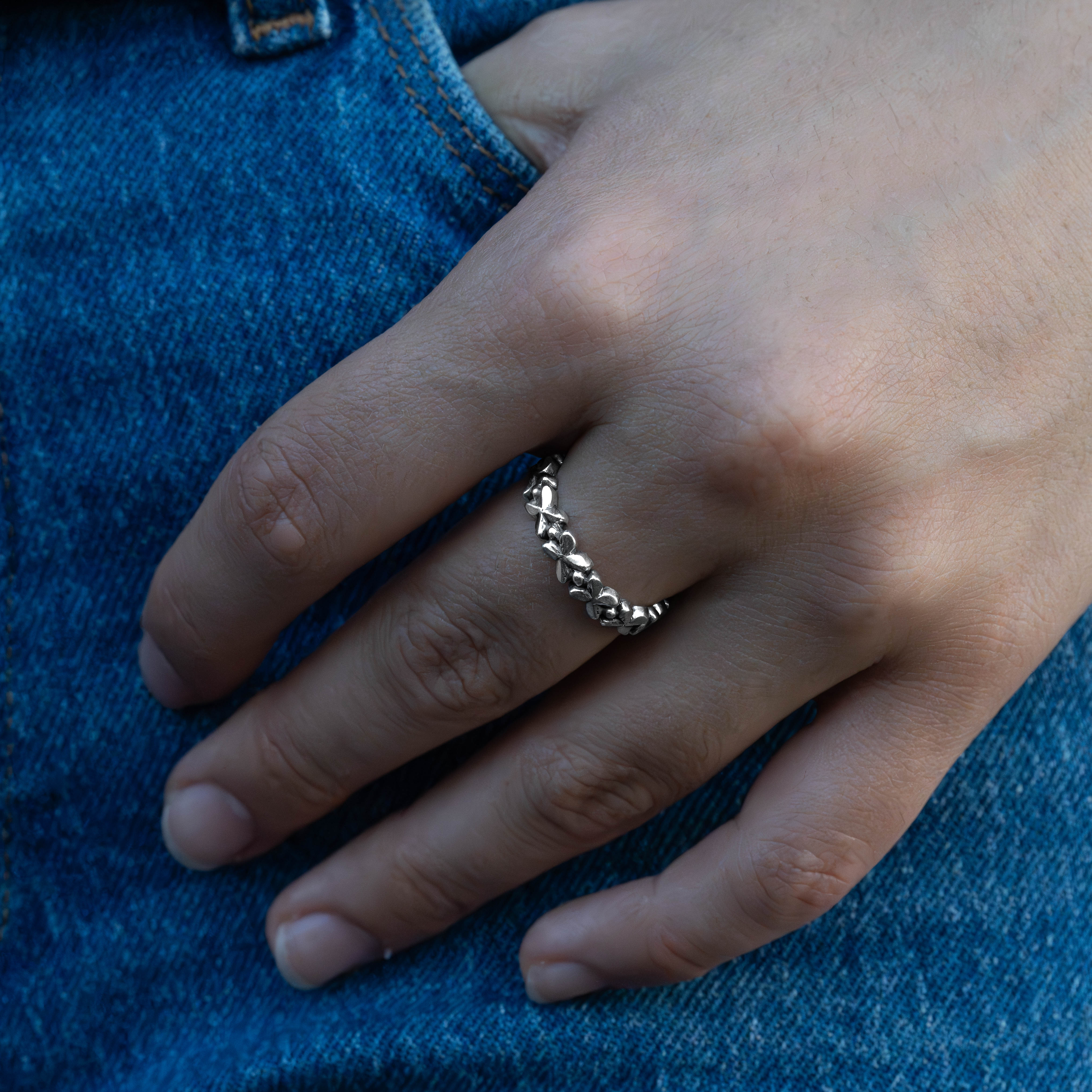A close-up of a hand wearing a thin silver ring with a textured, knotted bead-like design, resting on blue denim fabric.