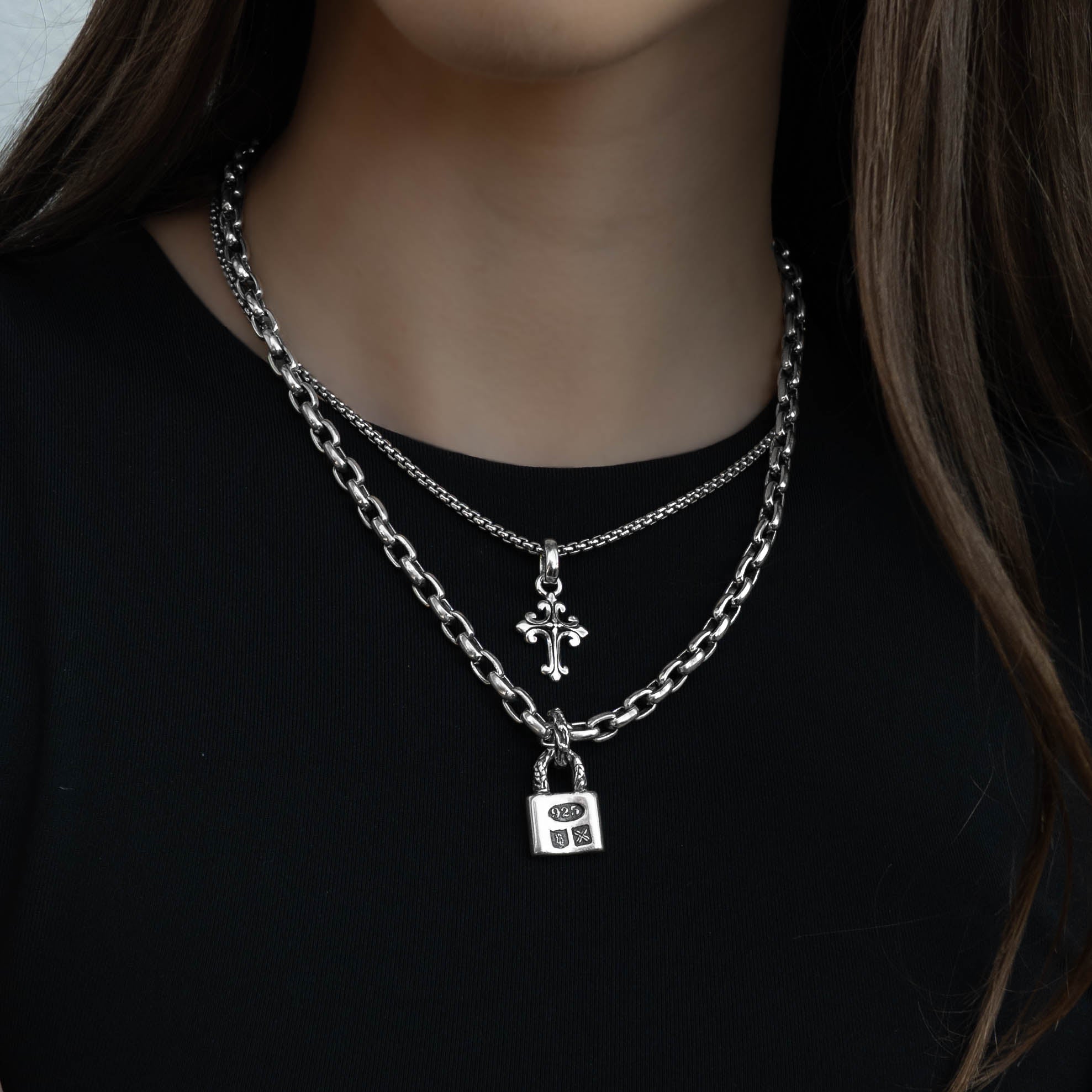 A close-up of a model wearing a layered silver necklace set. The set features a thick curb chain and a delicate chain with a small cross pendant and a padlock pendant.