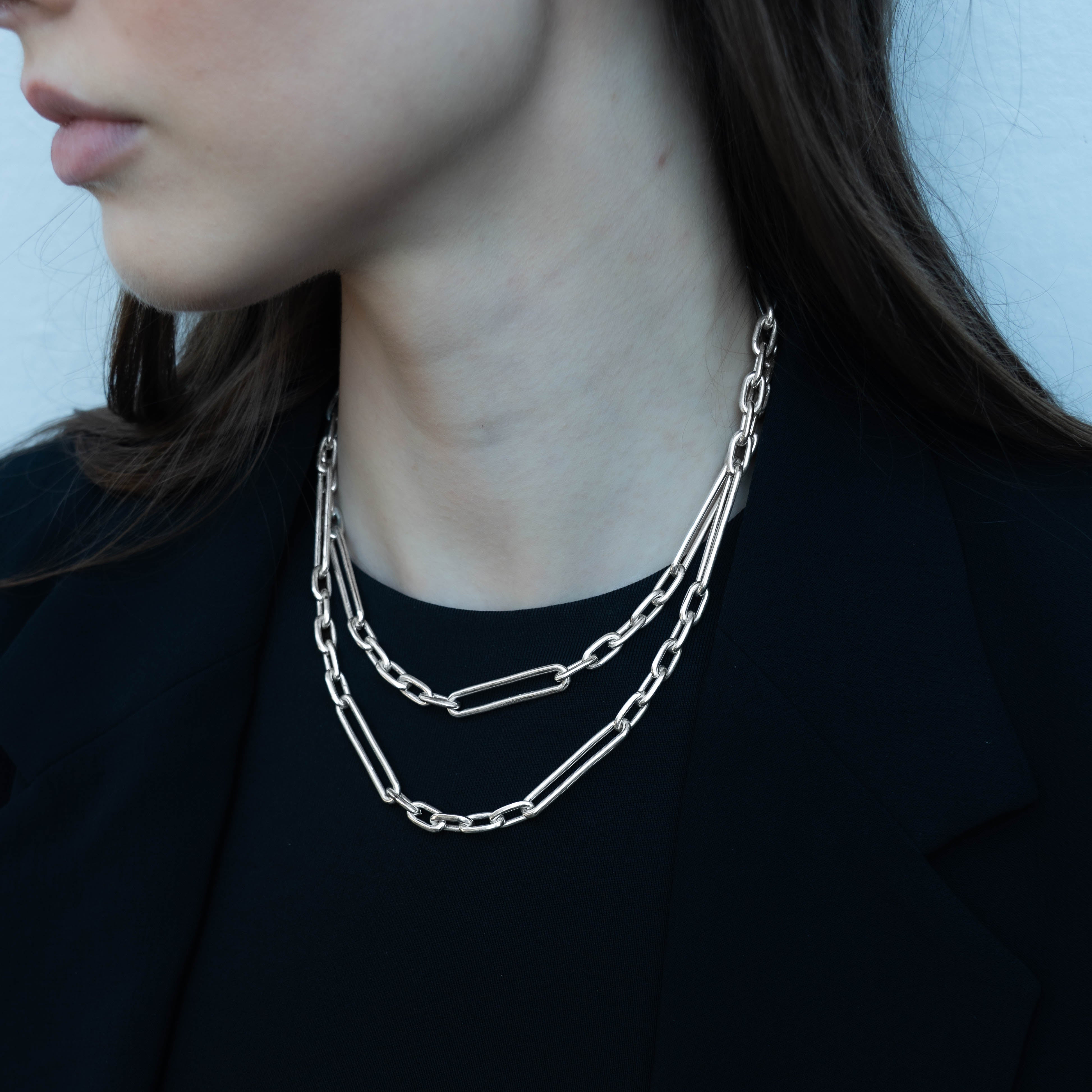 A close-up of a model wearing a layered silver chain necklace with paperclip and rectangular links over a black top.