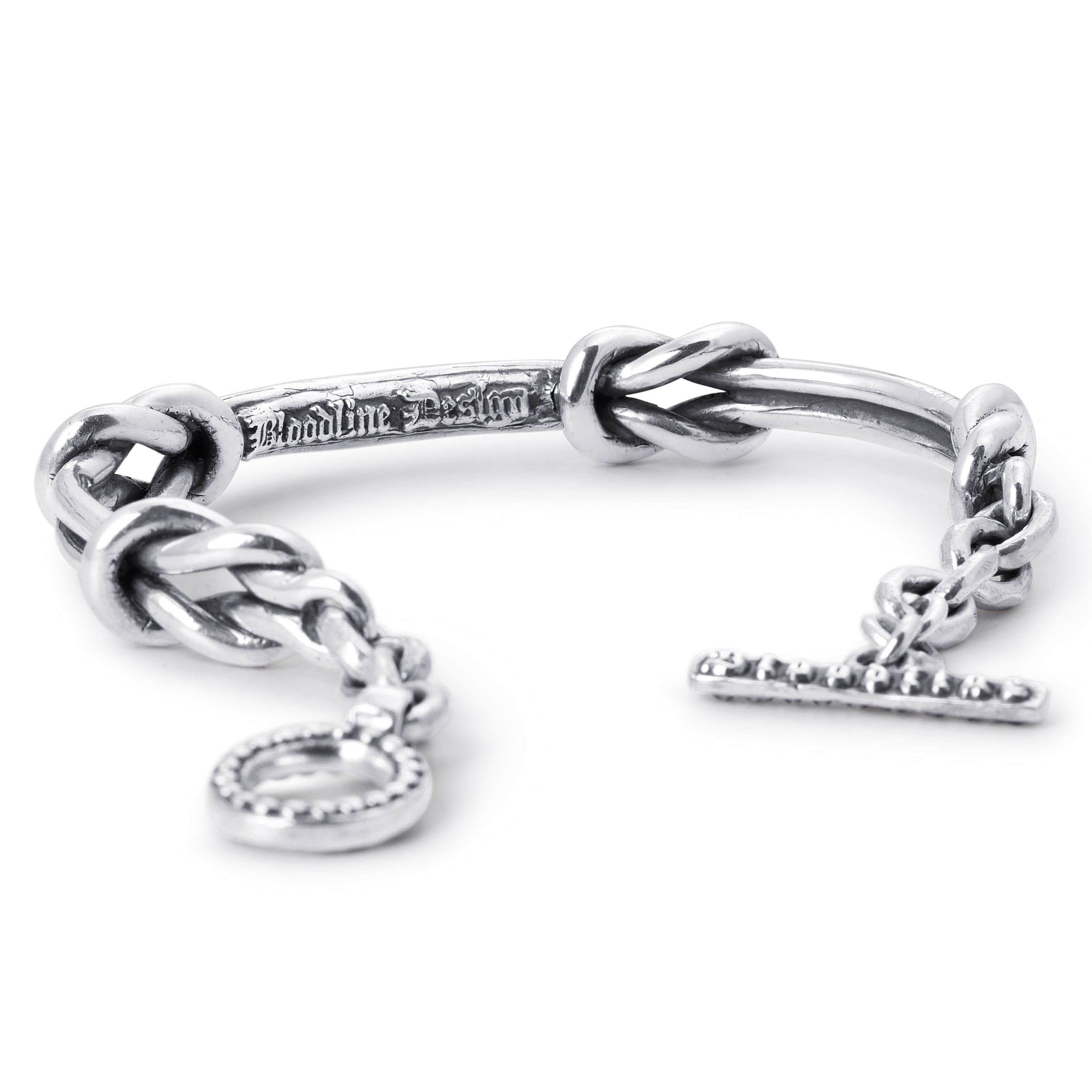 A silver-toned metal bracelet featuring a braided knot design, a short chain extension, a round bezel-set charm, and a T-bar clasp with engraved lettering, shown on a plain white background.