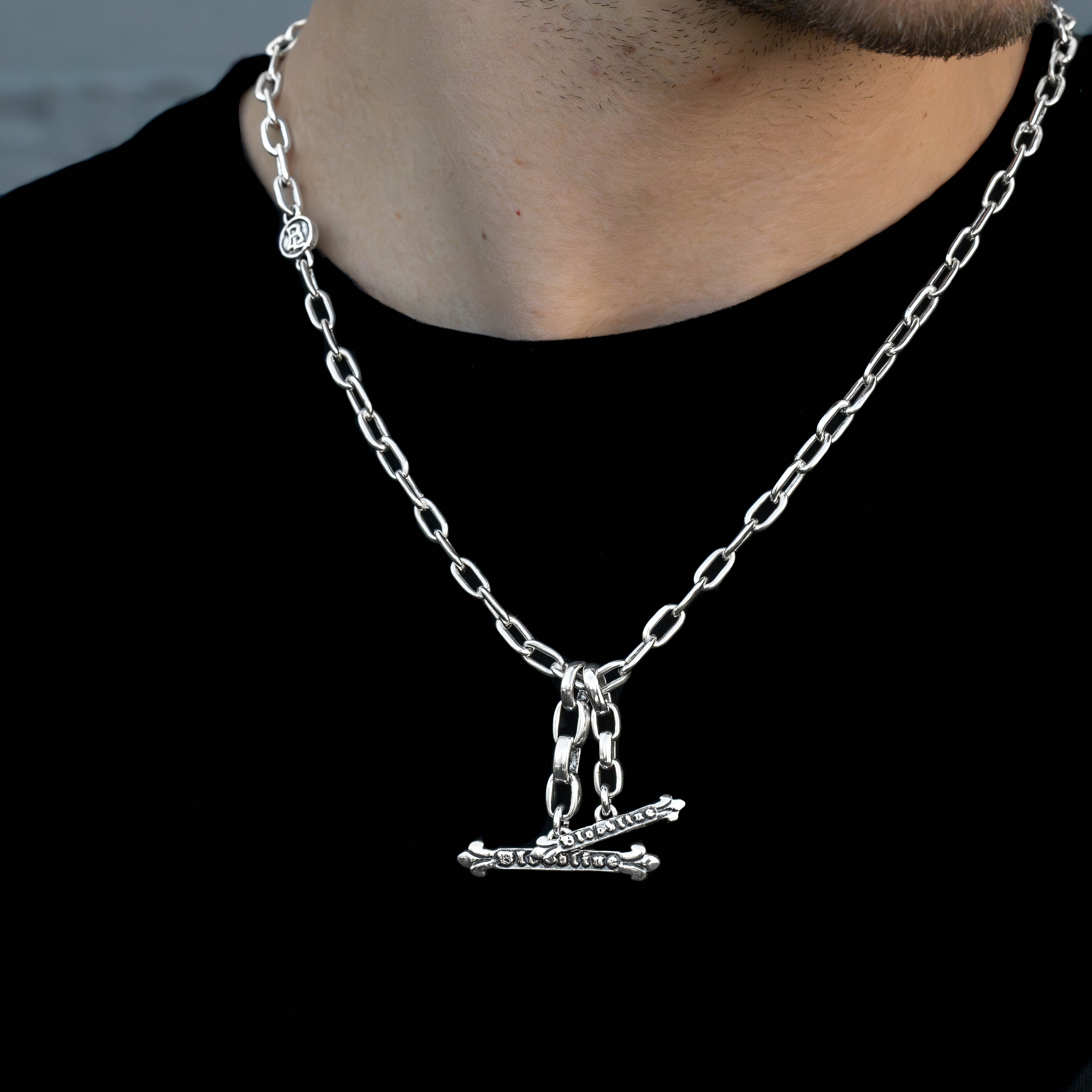 A close-up of a silver chain necklace worn by a person, featuring a horizontal T bar pendant set against a black shirt.