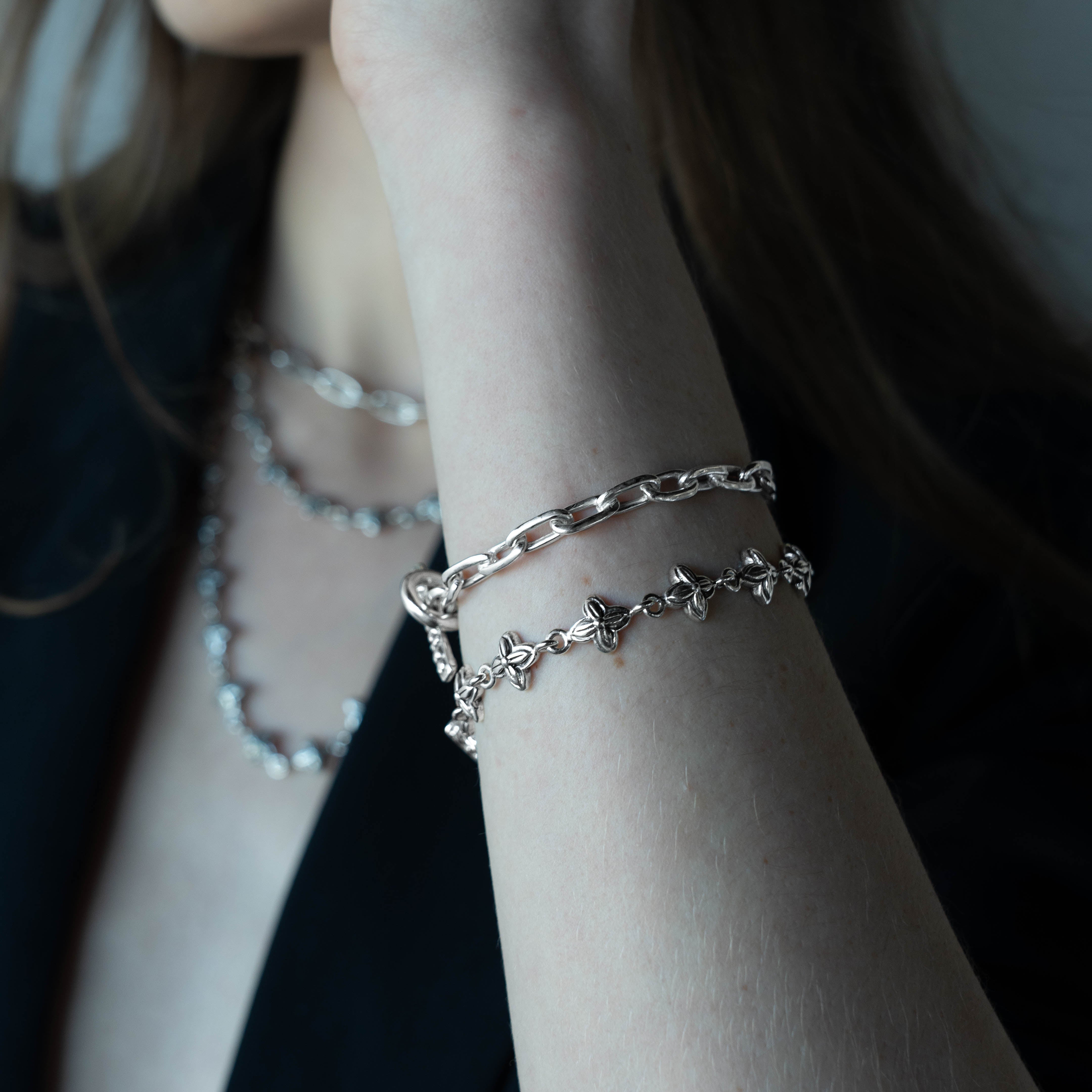 A close-up of a person's wrist wearing a layered silver bracelet set, featuring a thick curb chain and a delicate chain with small star charms, against a dark background.