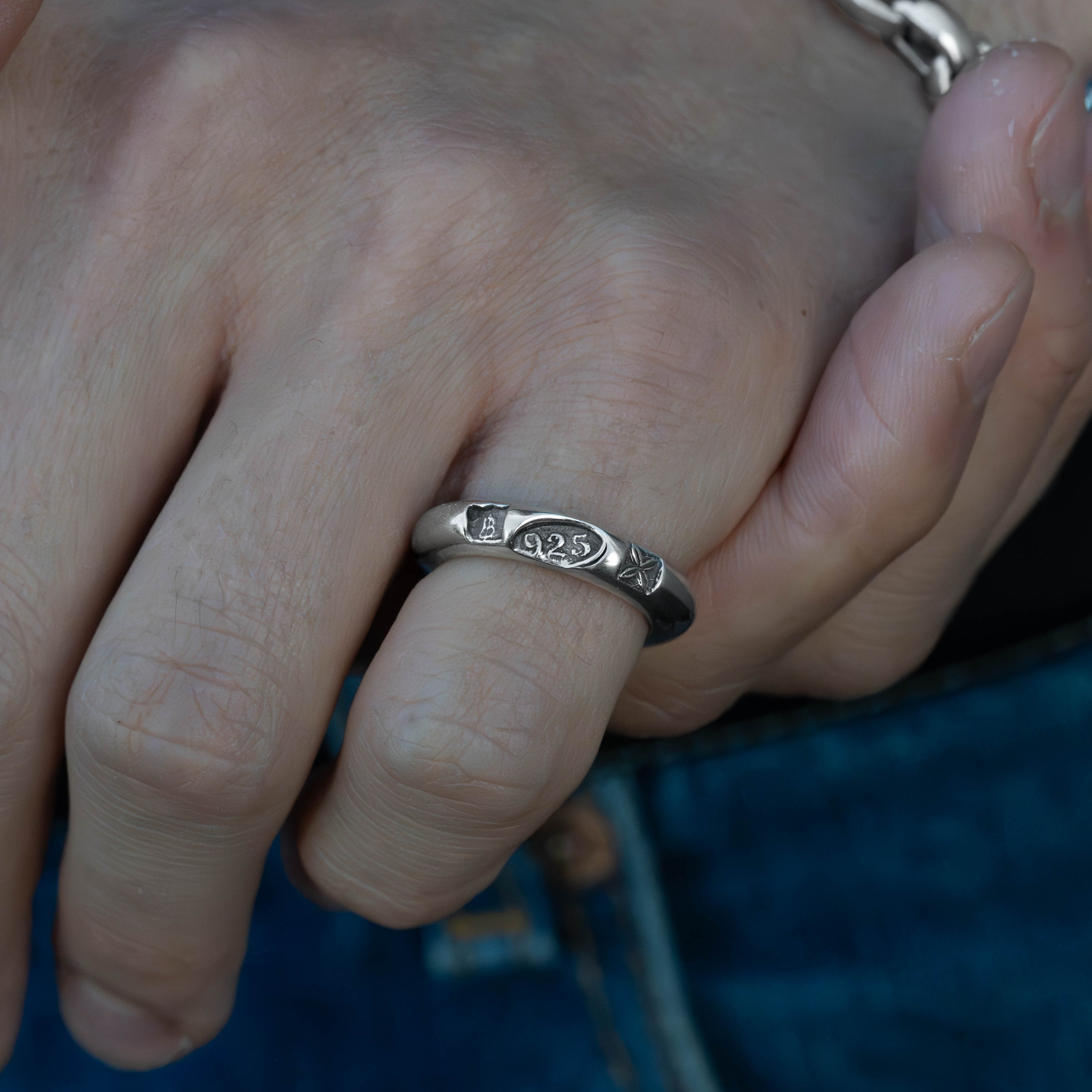 A close-up of a person's hand wearing a silver band ring with a small engraved symbol, with a silver chain bracelet on the wrist and blue jeans in the background.