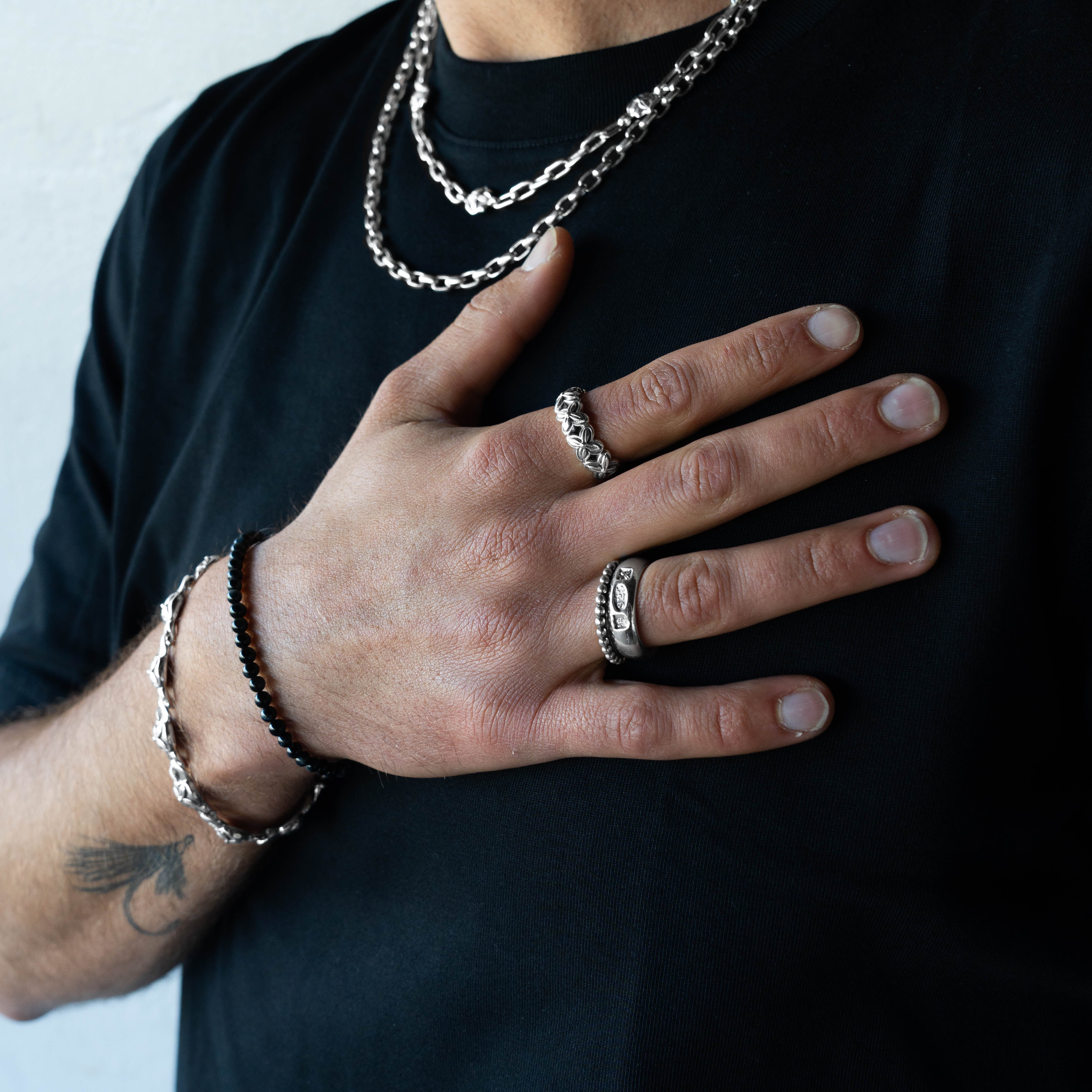 A close-up of a person's hand wearing multiple silver-toned jewelry pieces, including a textured ring, a chain necklace, and a beaded bracelet, over a black t-shirt.